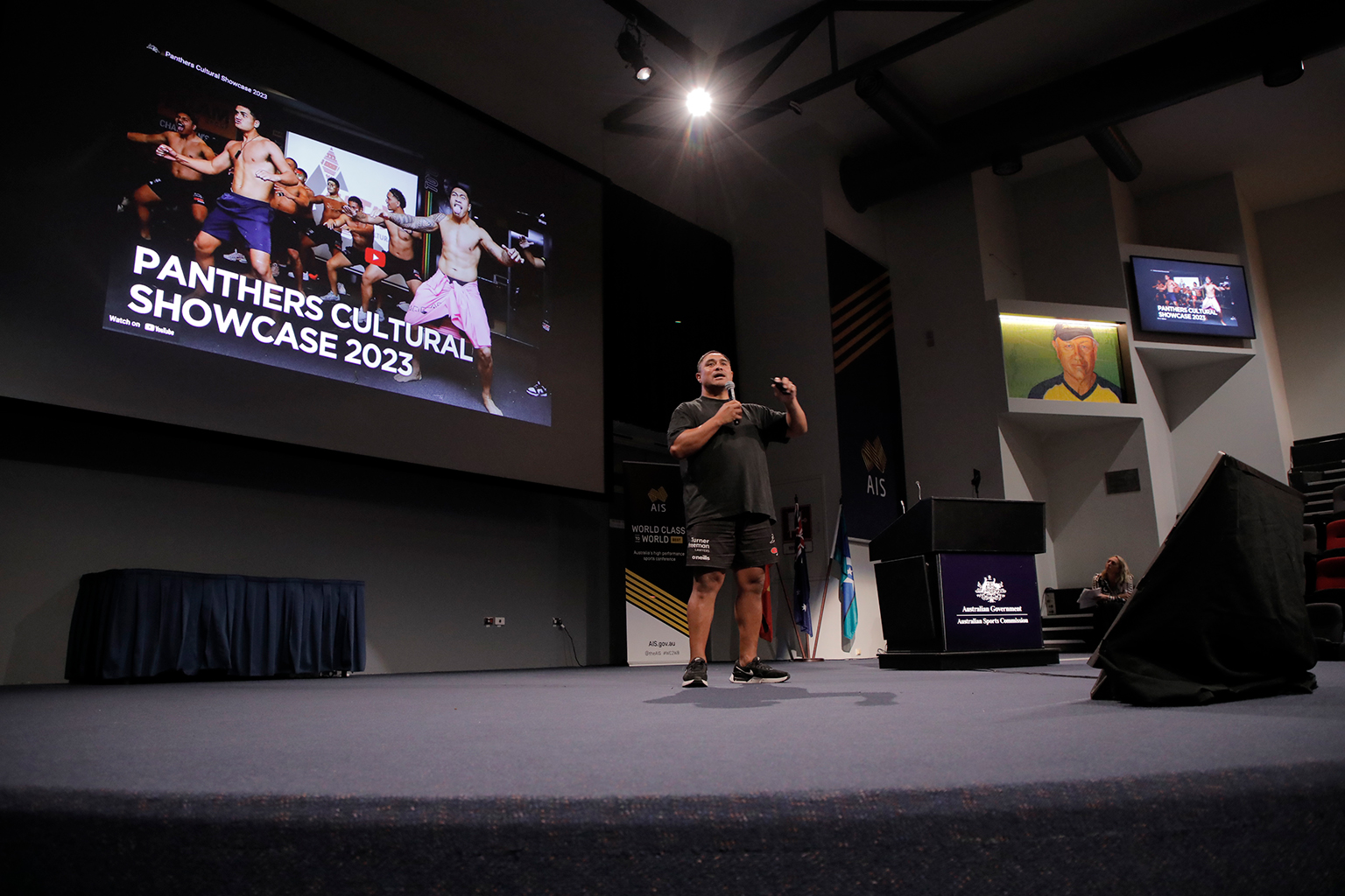 Wide image of a man on stage with a large projection screen in the background