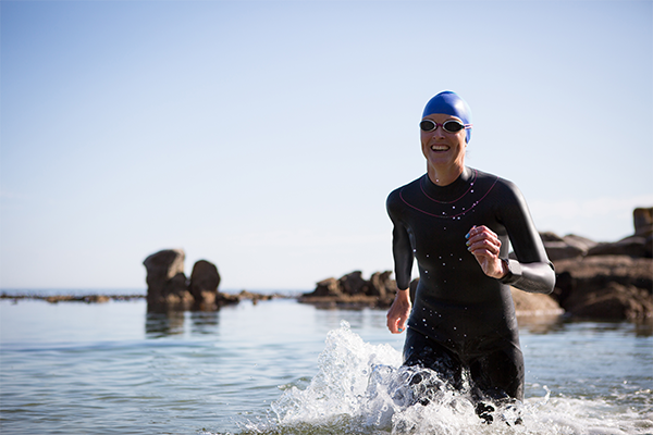 Person in a wetsuit, swim cap and goggles running out of calm open water near a rocky shoreline
