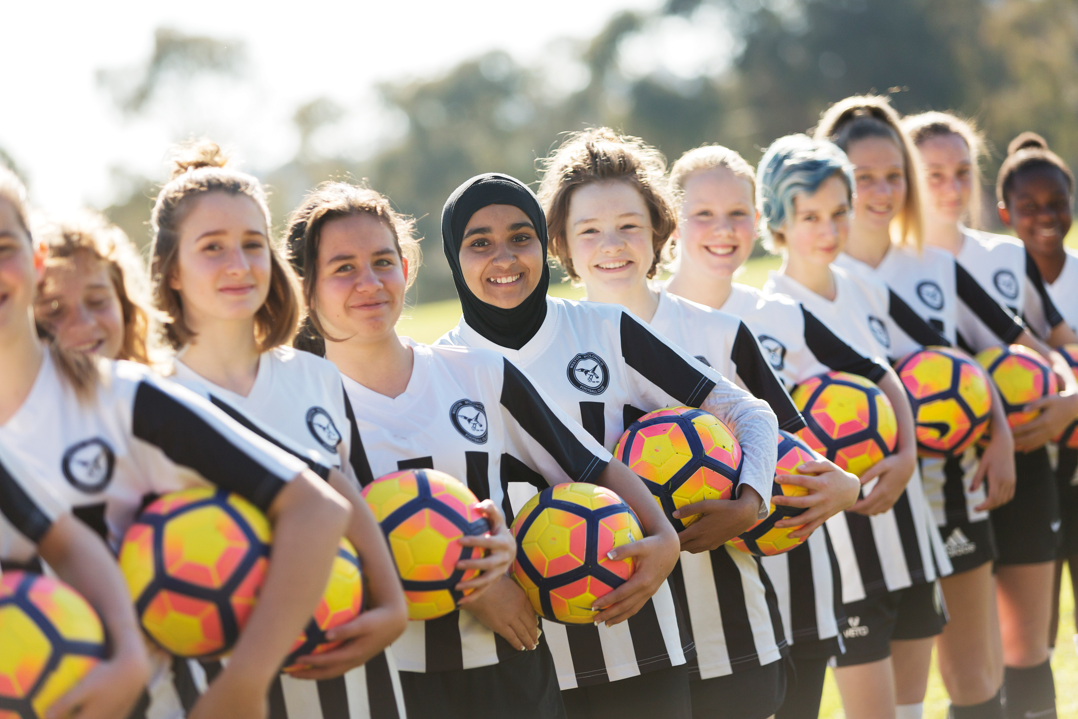 11 girls standing in line holding soccer balls in their football uniforms.