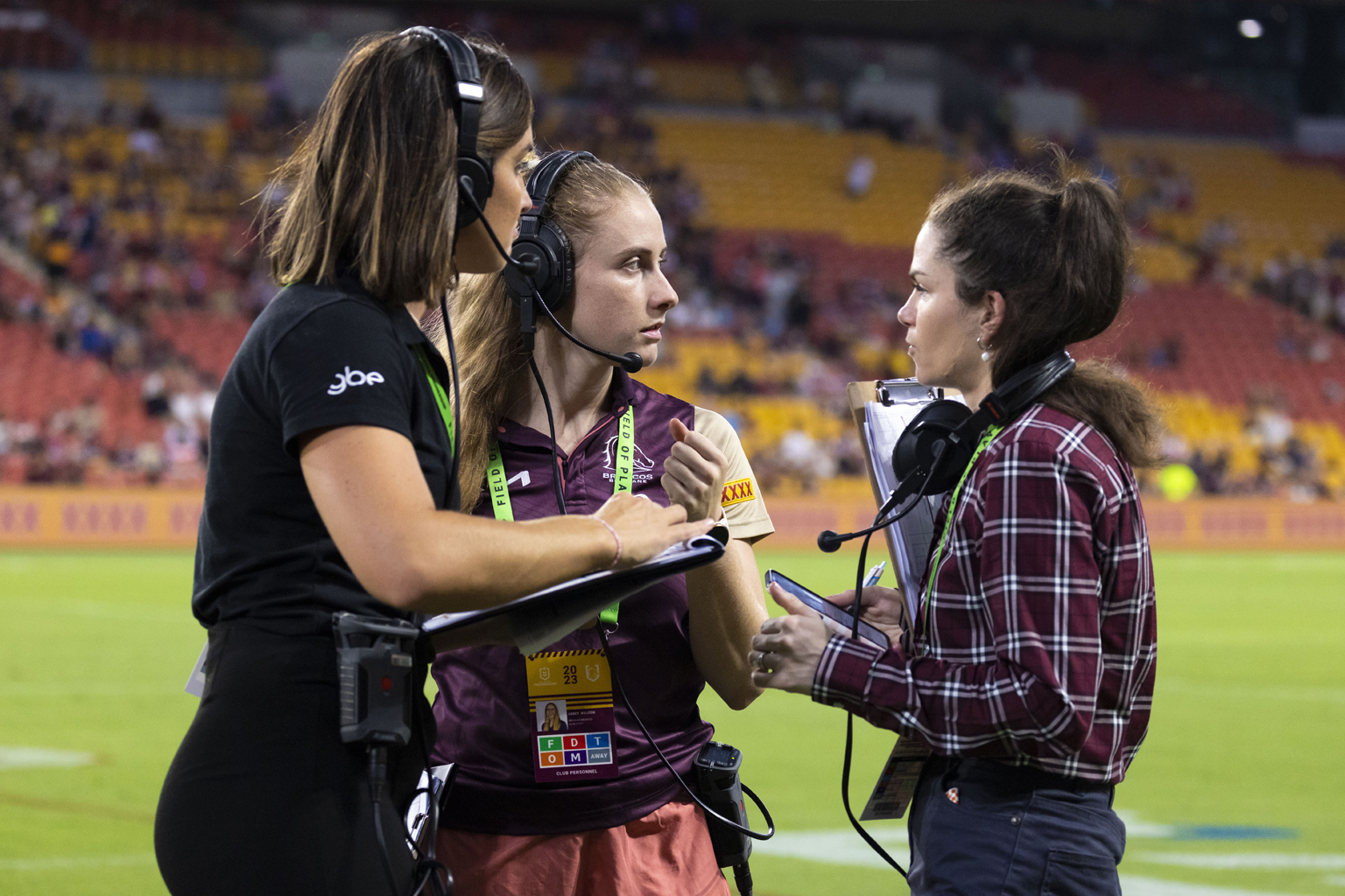 Three women on a sports field wearing communication headsets and carrying clipboards.