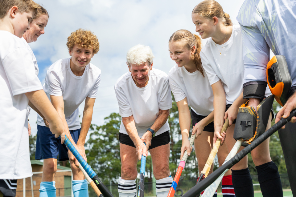 Seven people of varying ages standing in a circle wearing white t-shirts and holding hockey sticks