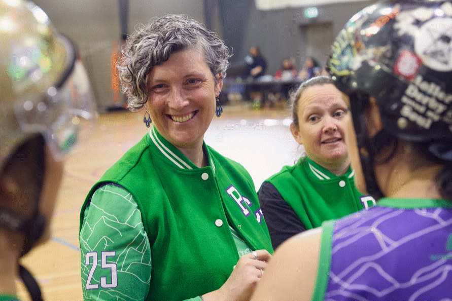 The image features four women from a roller derby team, two women are wearing vibrant green and looking at the camera, two others are standing in helmets with their backs to the camera.