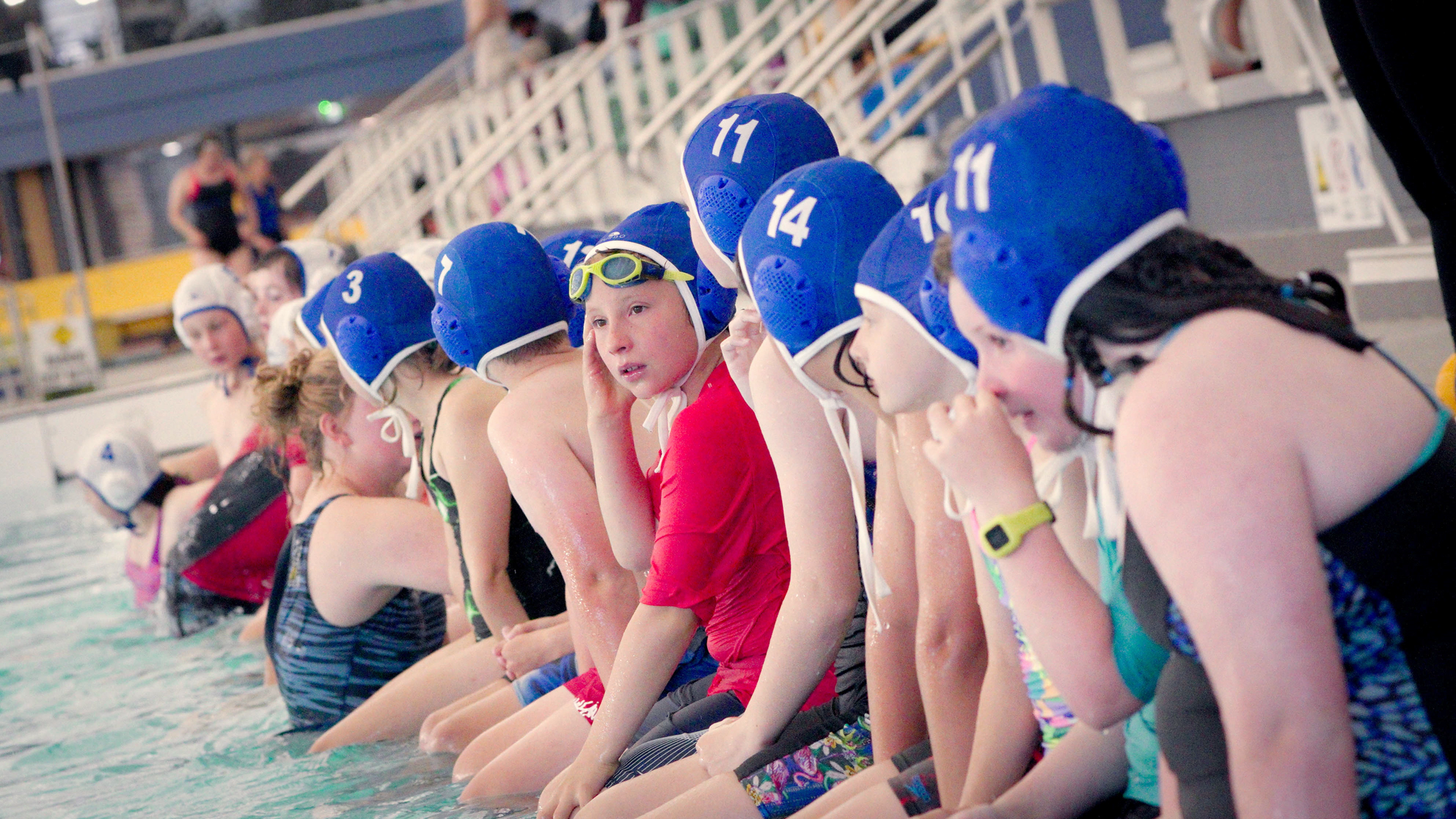 Children sitting on edge of pool with Water Polo caps on.