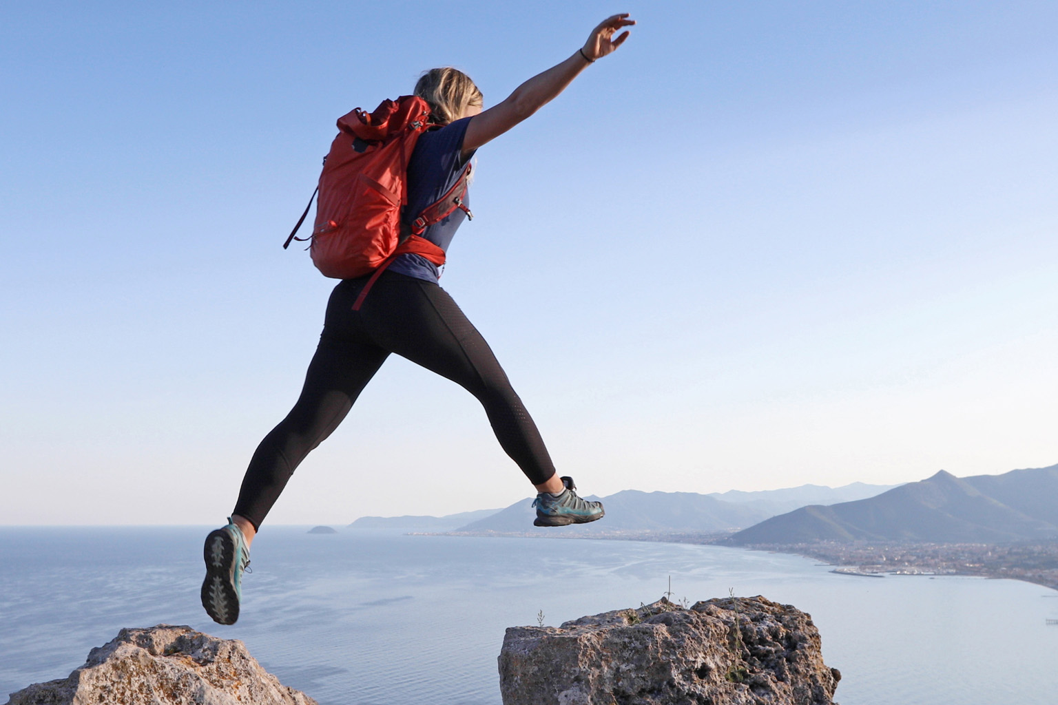 A woman wearing a hiking backpack leaps between to rocks with coastline and mountains in the background.