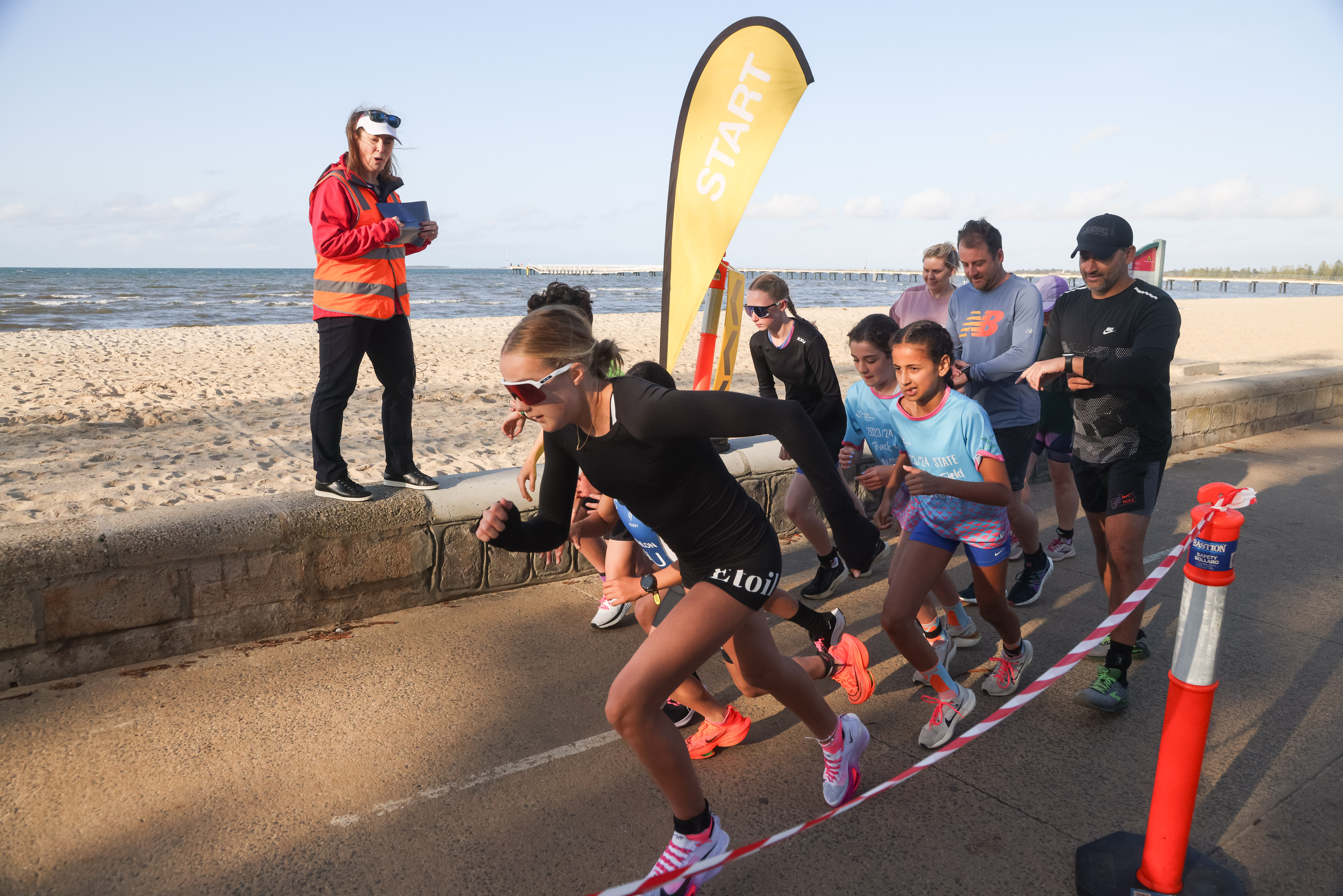 A triathlon official is starting a race with young participants beginning to run. There is a yellow tear drop flag saying 'start.'