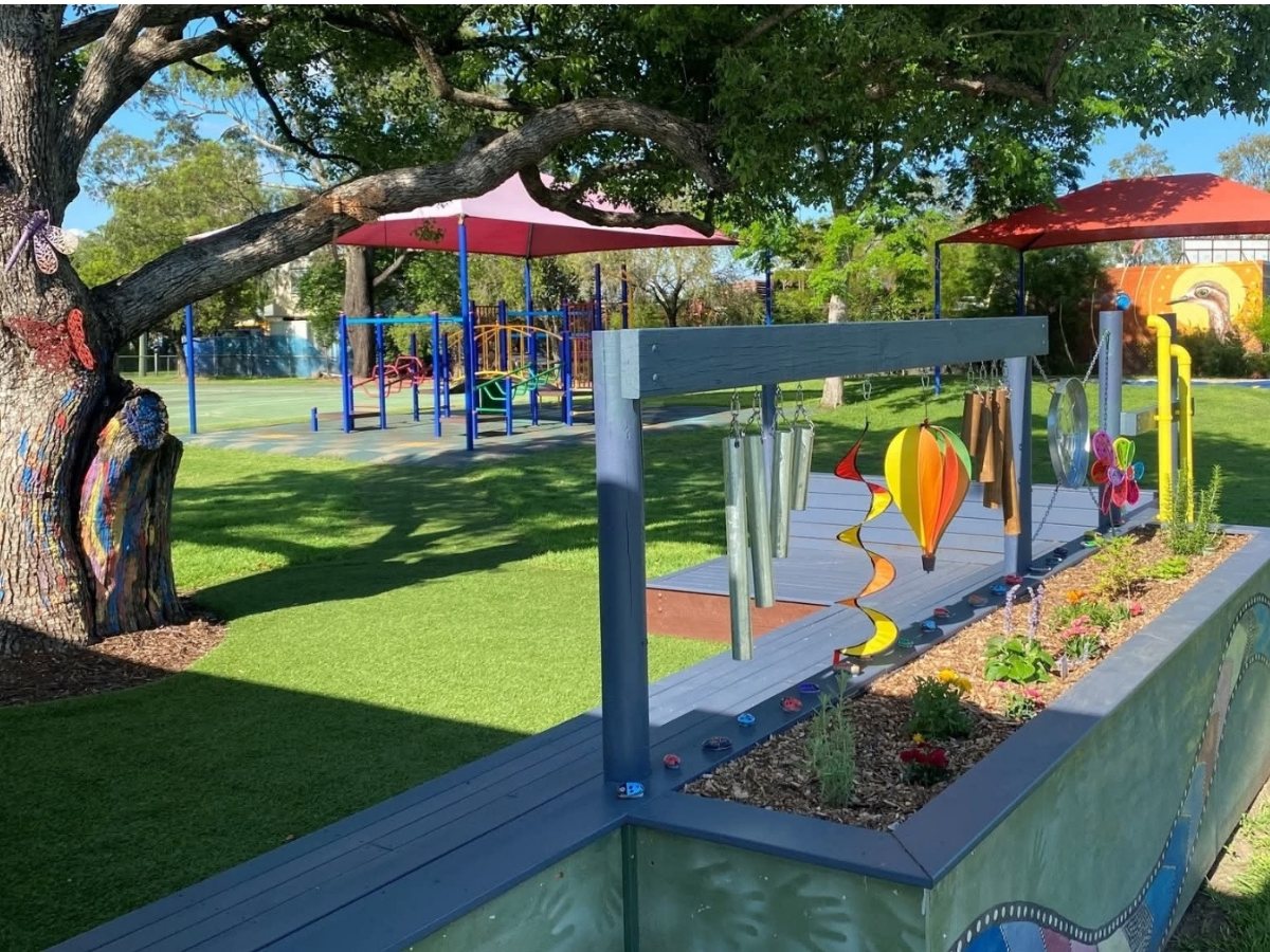 A photo of Rocklea State School that is popping with colour, wind chimes, bright green grass and big trees hanging over a veggie patch