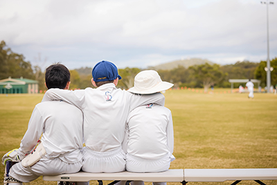 Three boys sitting on a bench during a cricket match