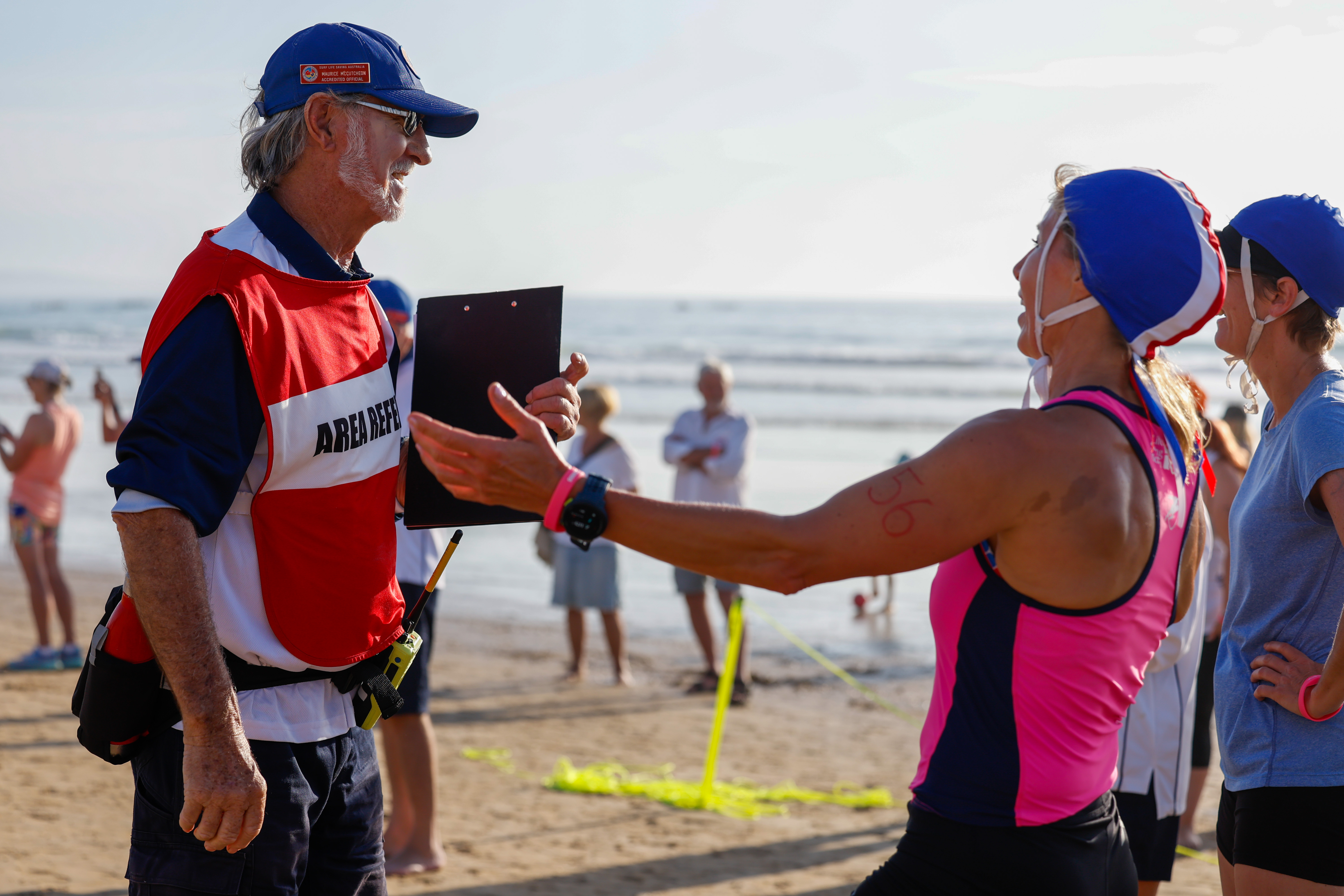 A surf lifesaving official is smiling and talking with some participants who are wearing caps.