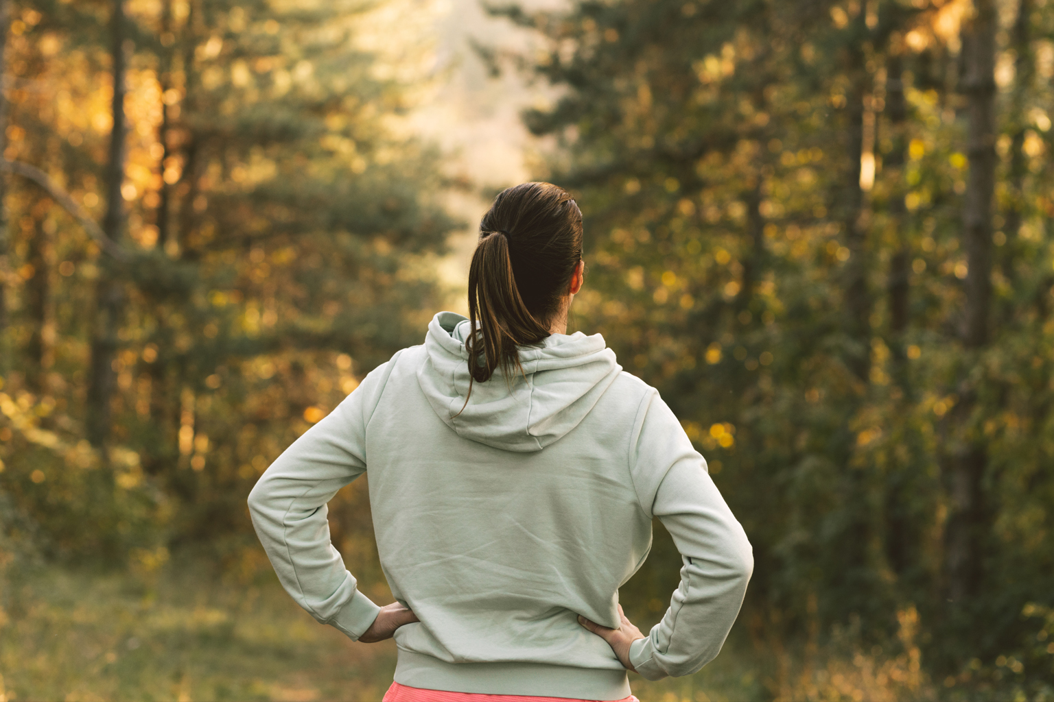 A woman in a hoodie looks out towards forested area with hands on hips.