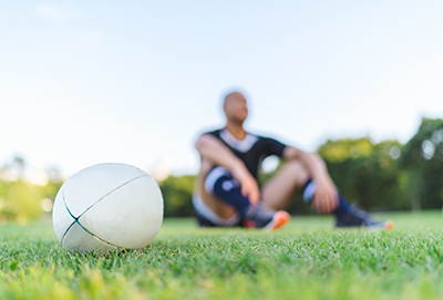 A rugby ball is put on grass field while a rugby player is sitting on grass in the background