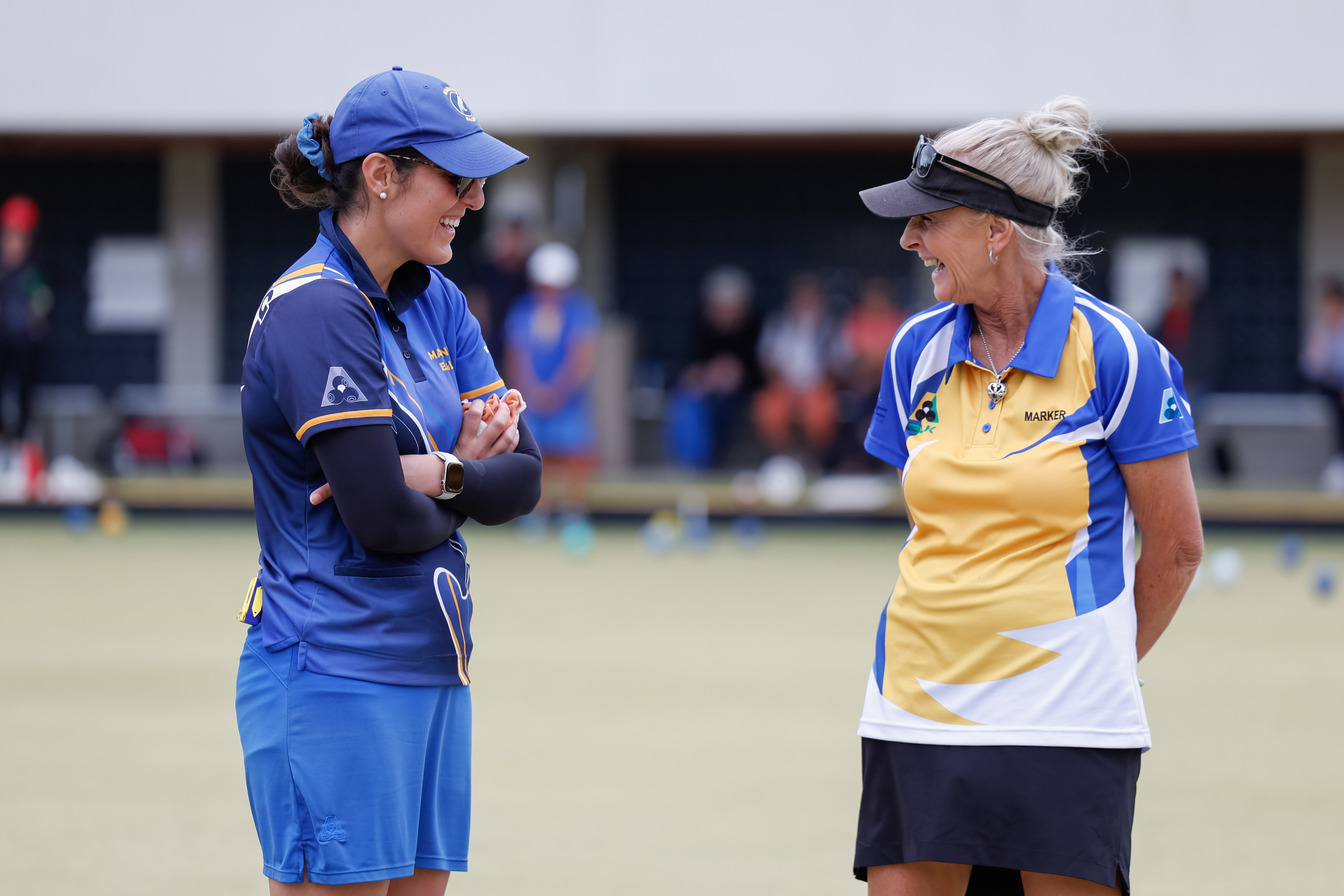 A lawn bowls official wearing a yellow shirt is standing on the green talking to a participant wearing a blue uniform.