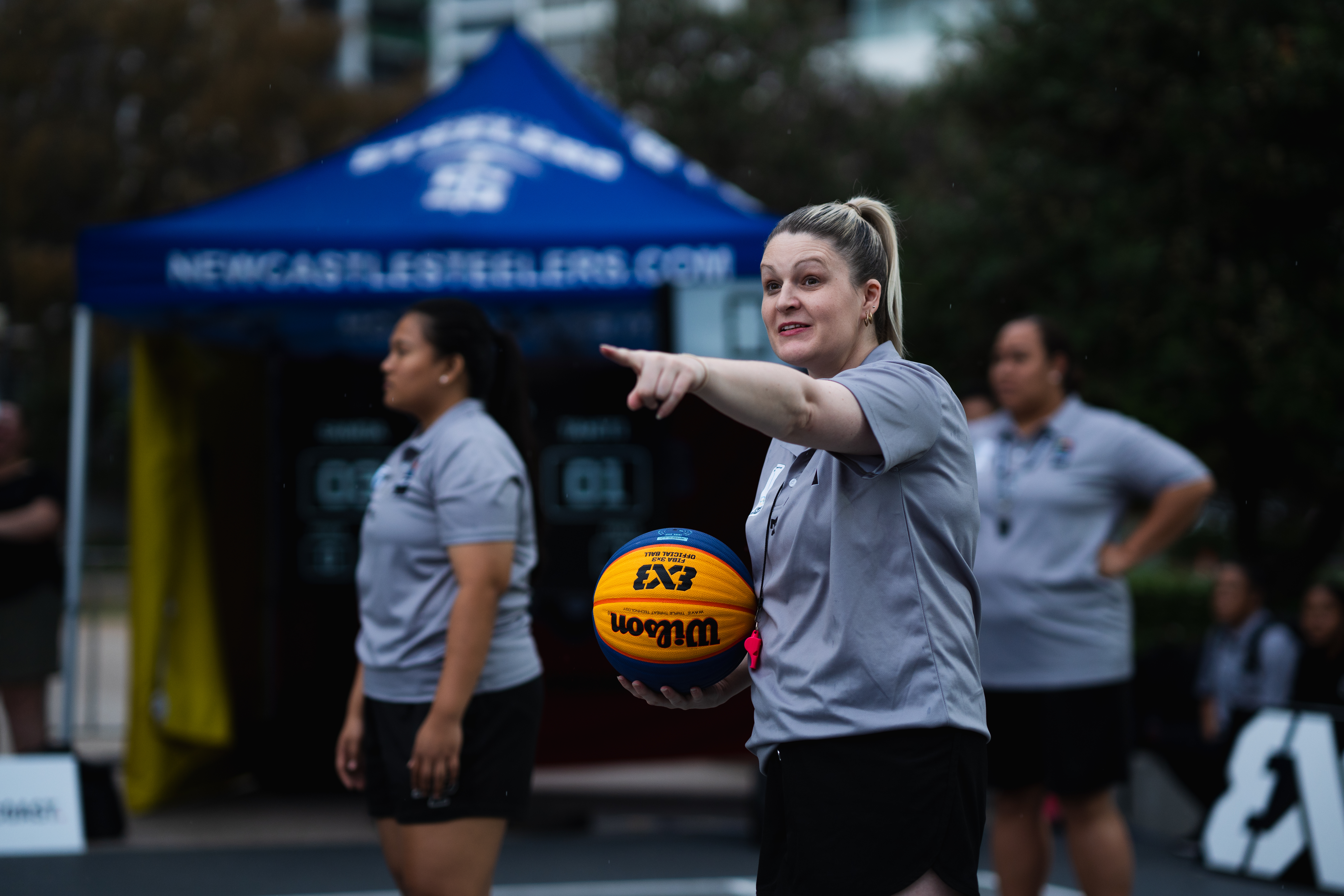 A basketball official holding a ball is smiling and pointing. There are two officials in the background.
