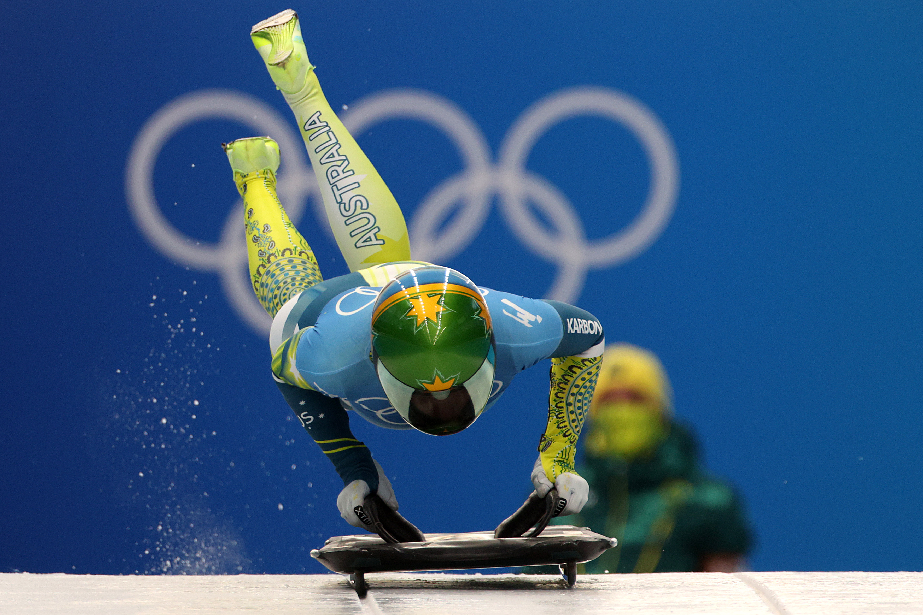 Image shows a skeleton racer jumping into their sled. The racer is wearing a green and gold skinsuit.