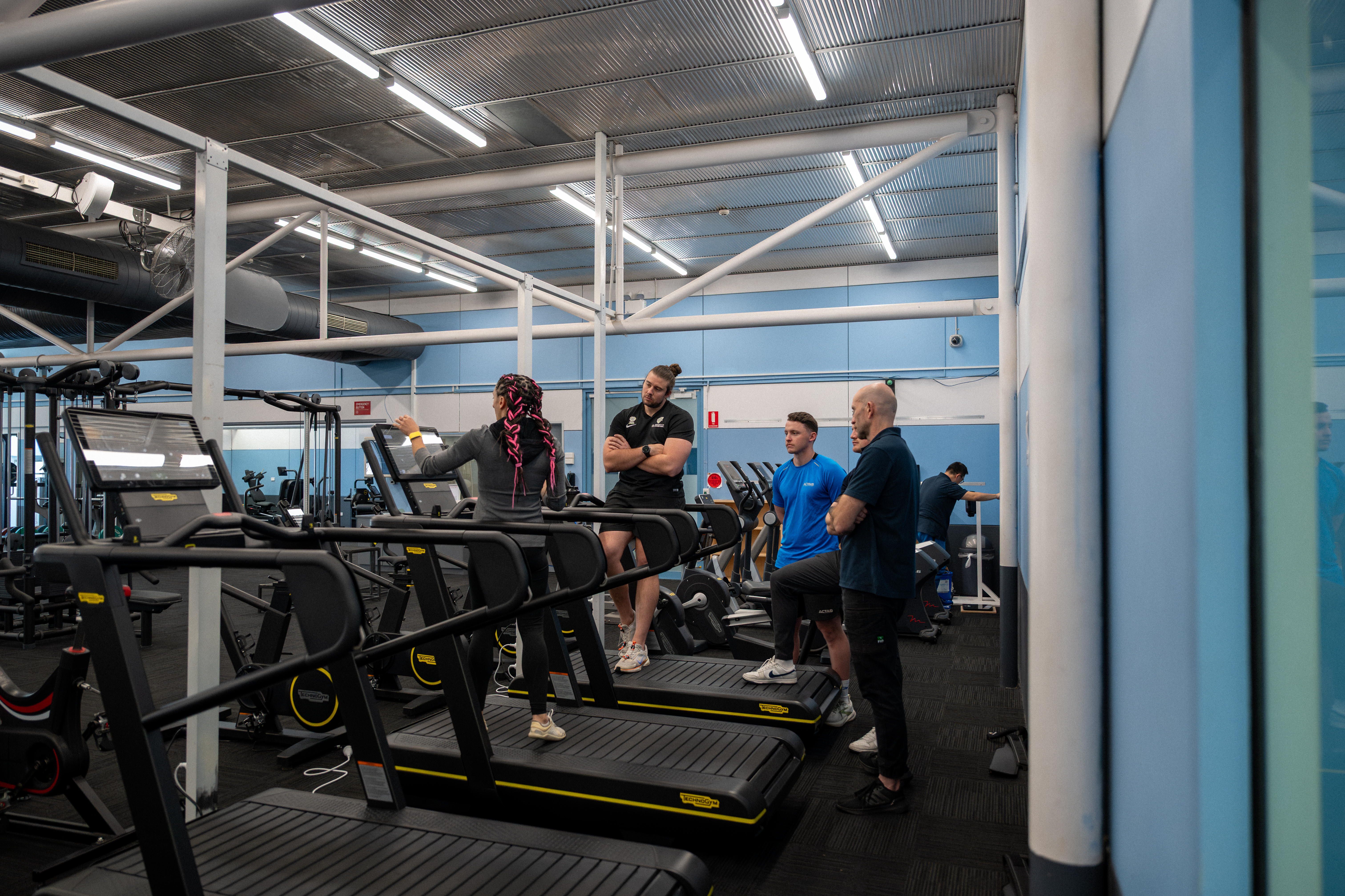 ASC staff watch a demonstration of a Technogym treadmill machine, which sits in a row of machines, inside the AIS Strength and Conditioning Gym