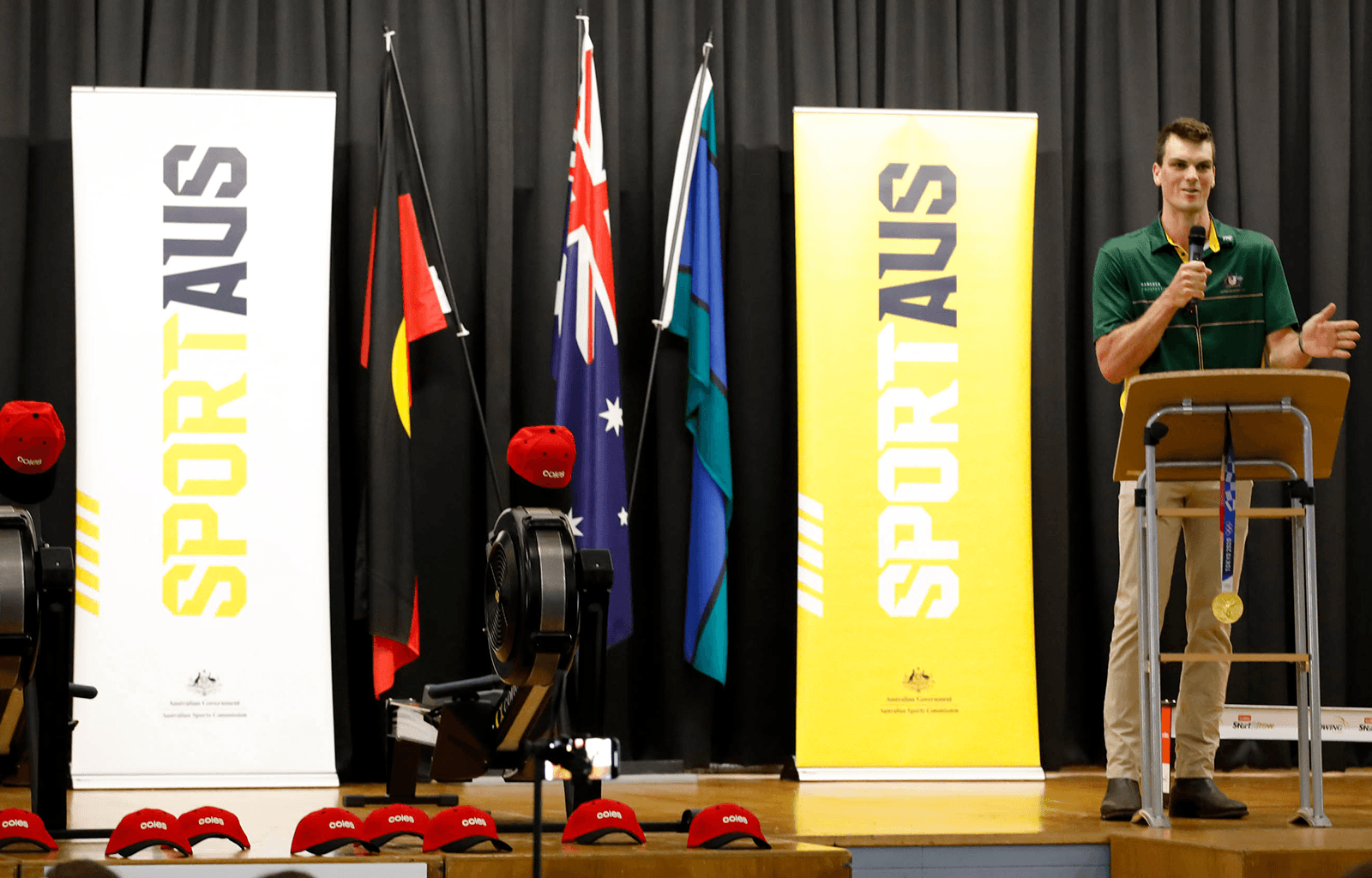Alex Purnell stands at a lectern on stage with SportAUS banners, rowing machines and Australian, Aboriginal and Torres Strait Island flags. Red Coles caps are spread across the stage.