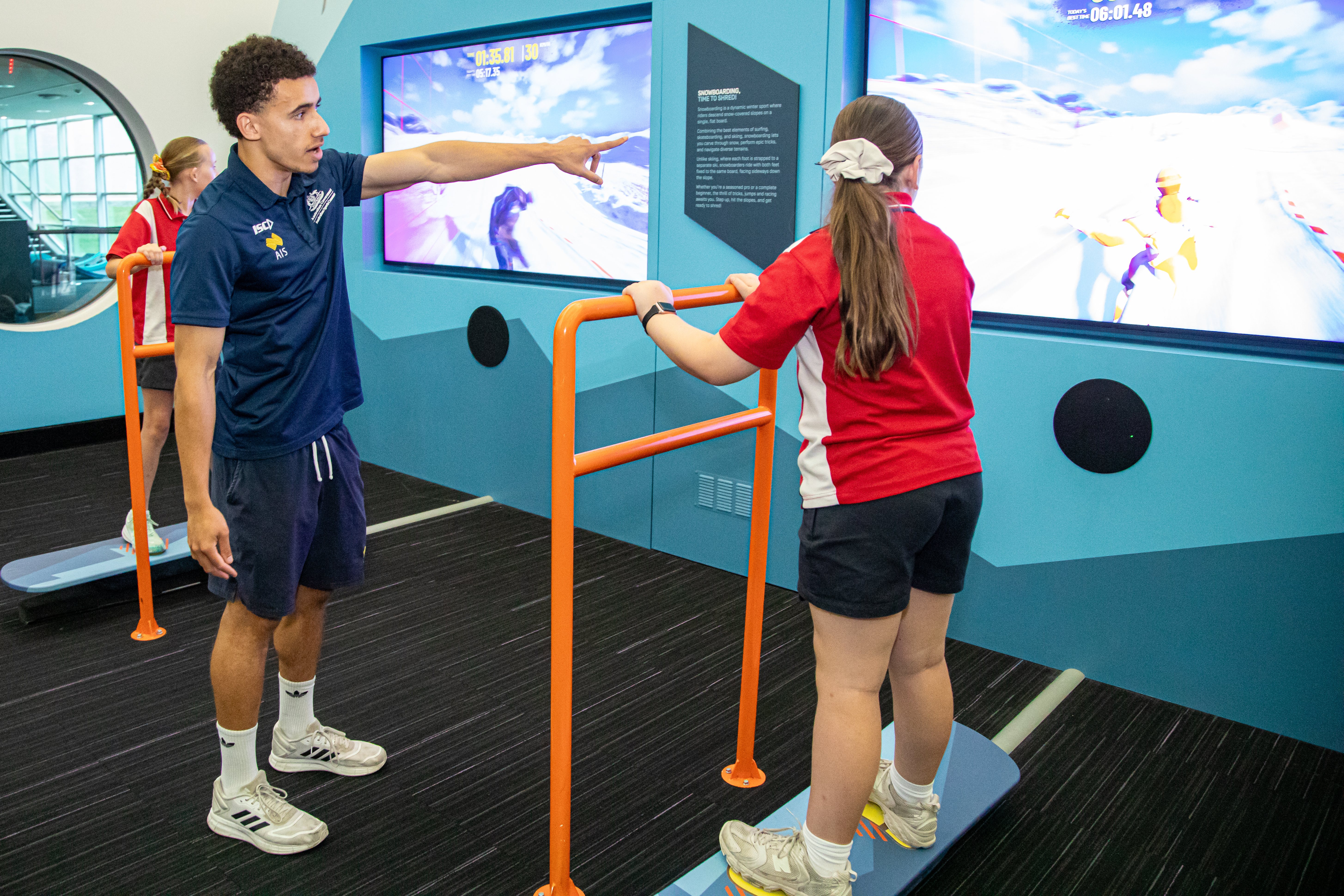 An image of two school children trying the new snowboarding interactive exhibit, helped by an AIS staff member.
