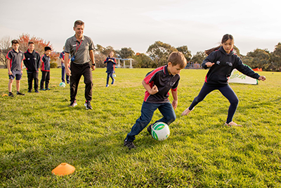 Boy dribbling a soccer ball with a girl defending. Coach watching.