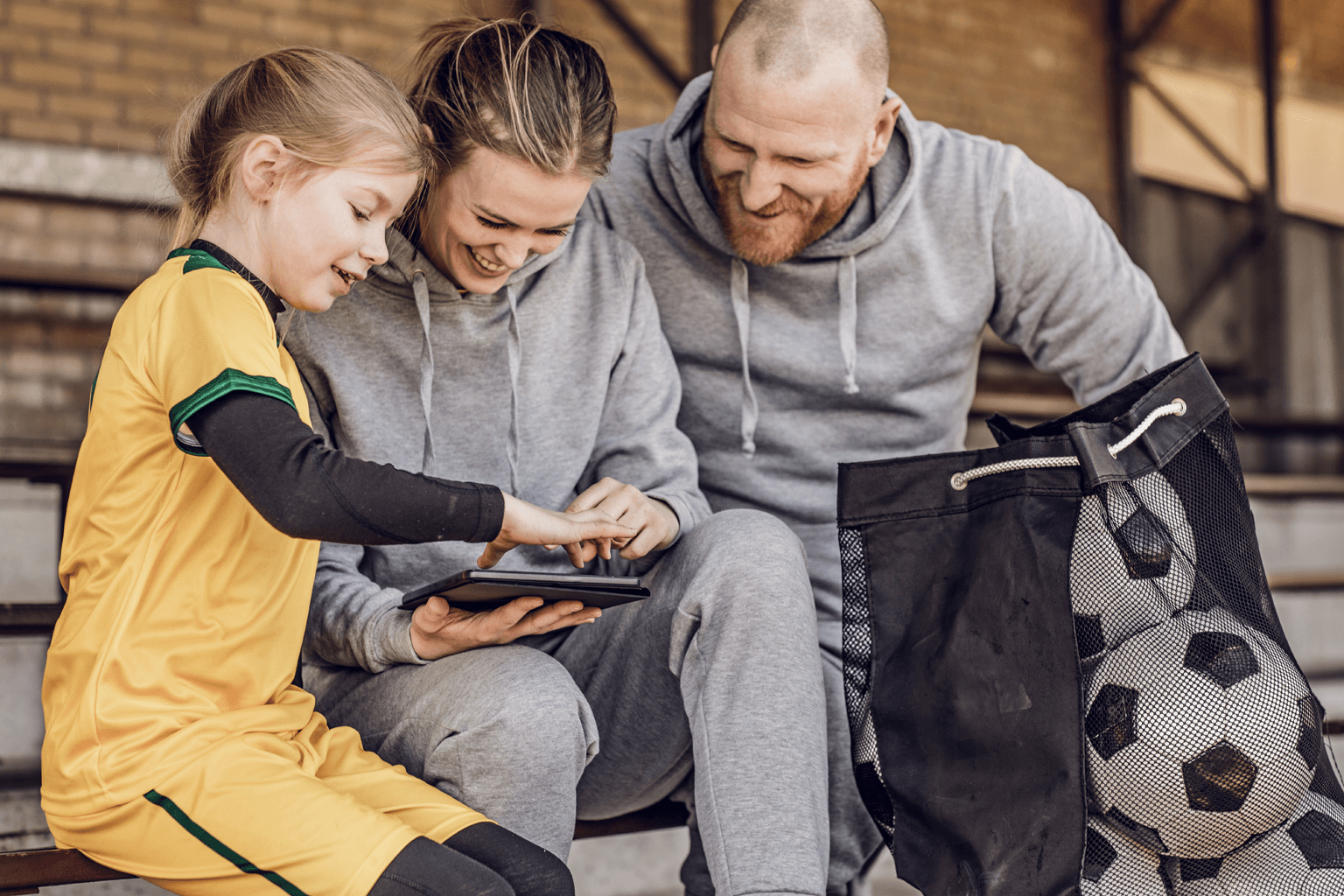 Girl sits with a woman an a man next to a bag of soccer balls, looking at an iPad