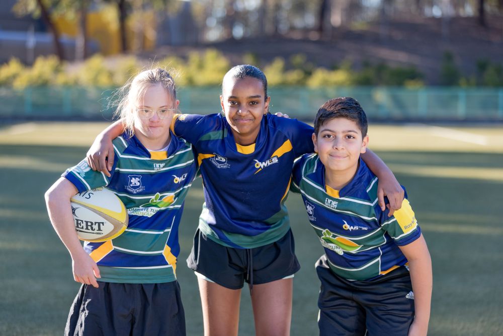 Three children wearing rugby uniforms in a huddle on a playing field facing towards the camera.