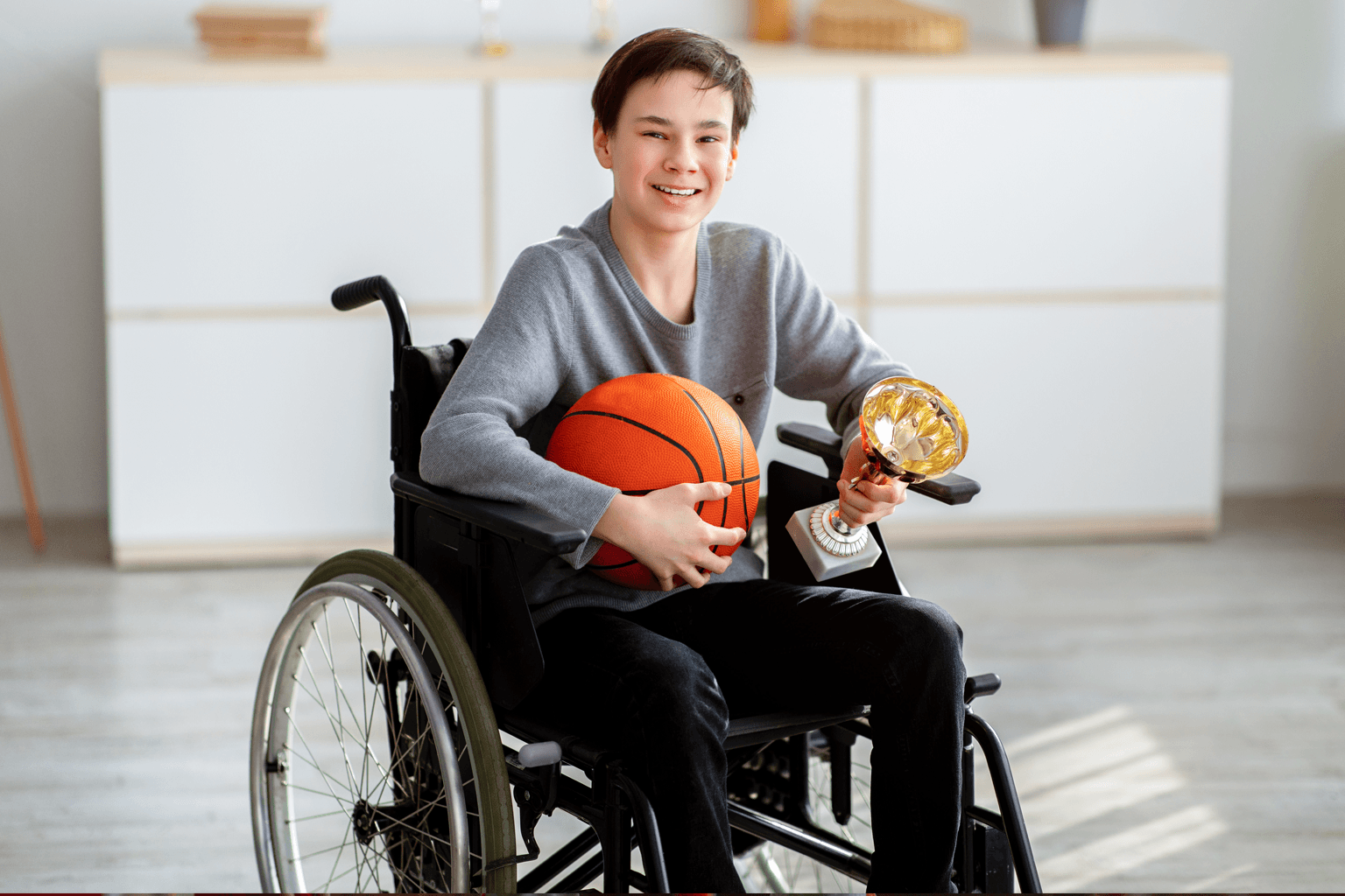 A boy sits in a wheelchair holding a basketball in one hand and a trophy in the other.
