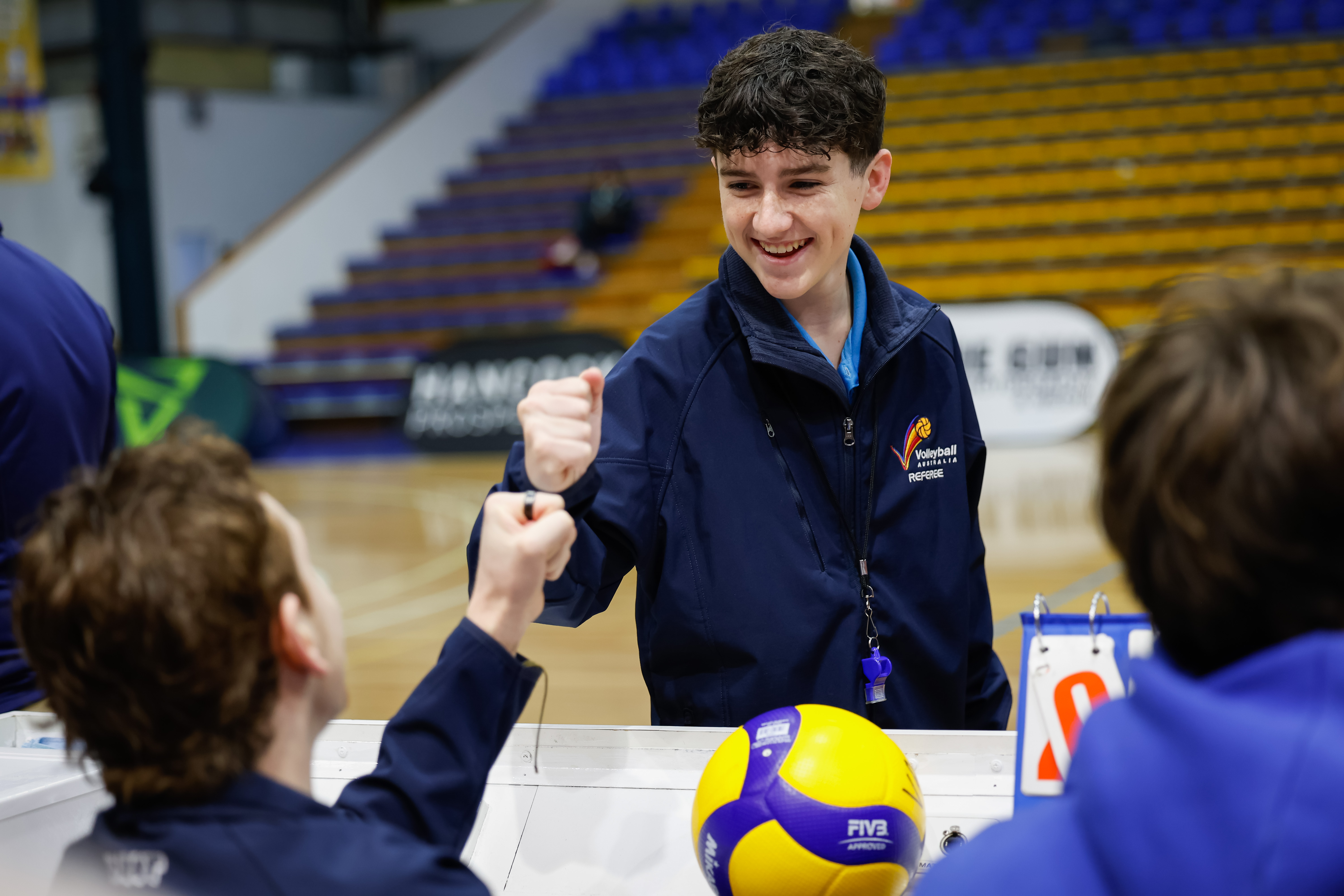 A young volleyball official wearing a navy blue jacket fist pumps another official. There is a volleyball on the table.