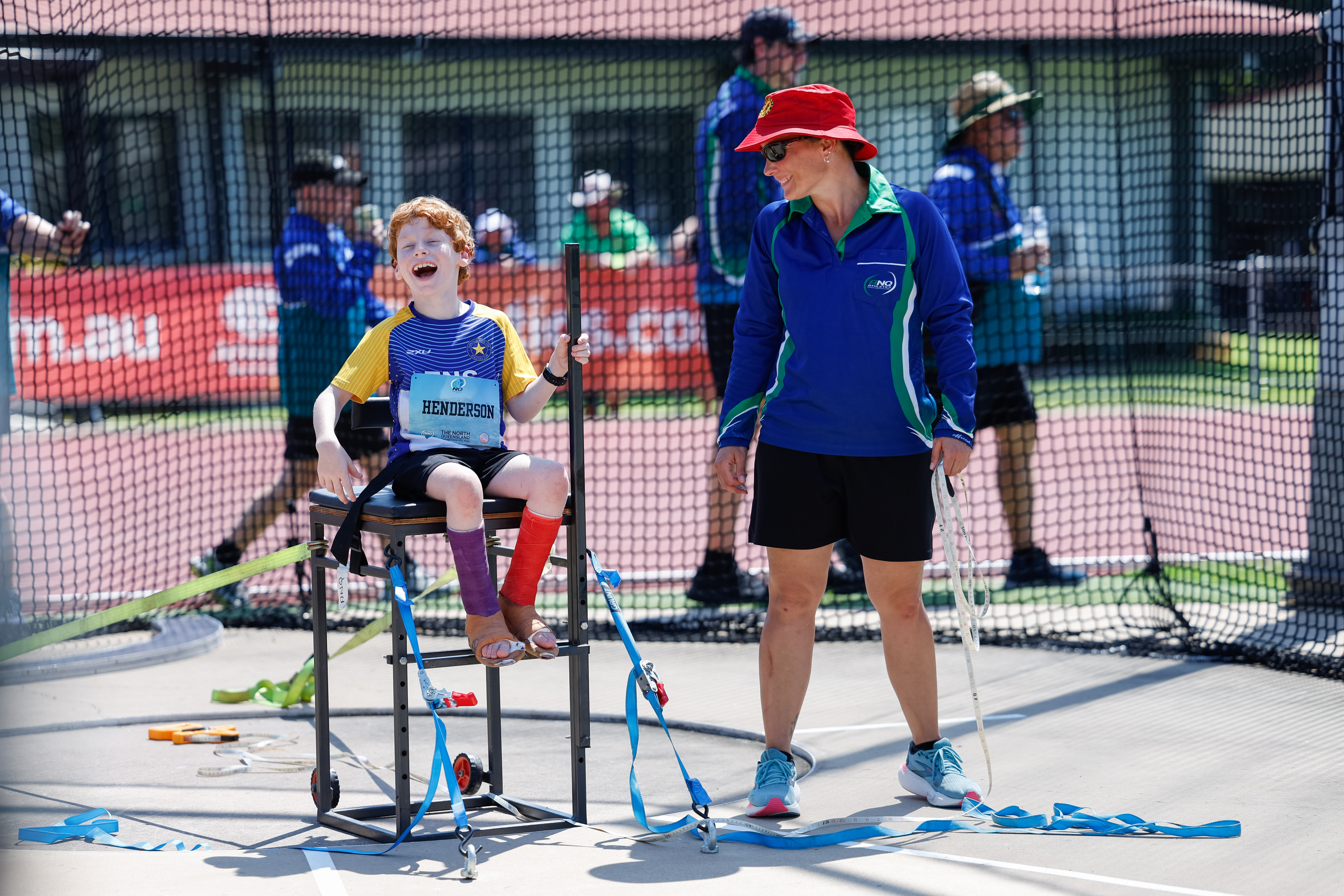 An athletics official is standing next to and smiling at a young participant who is sitting in a throwing chair and laughing.