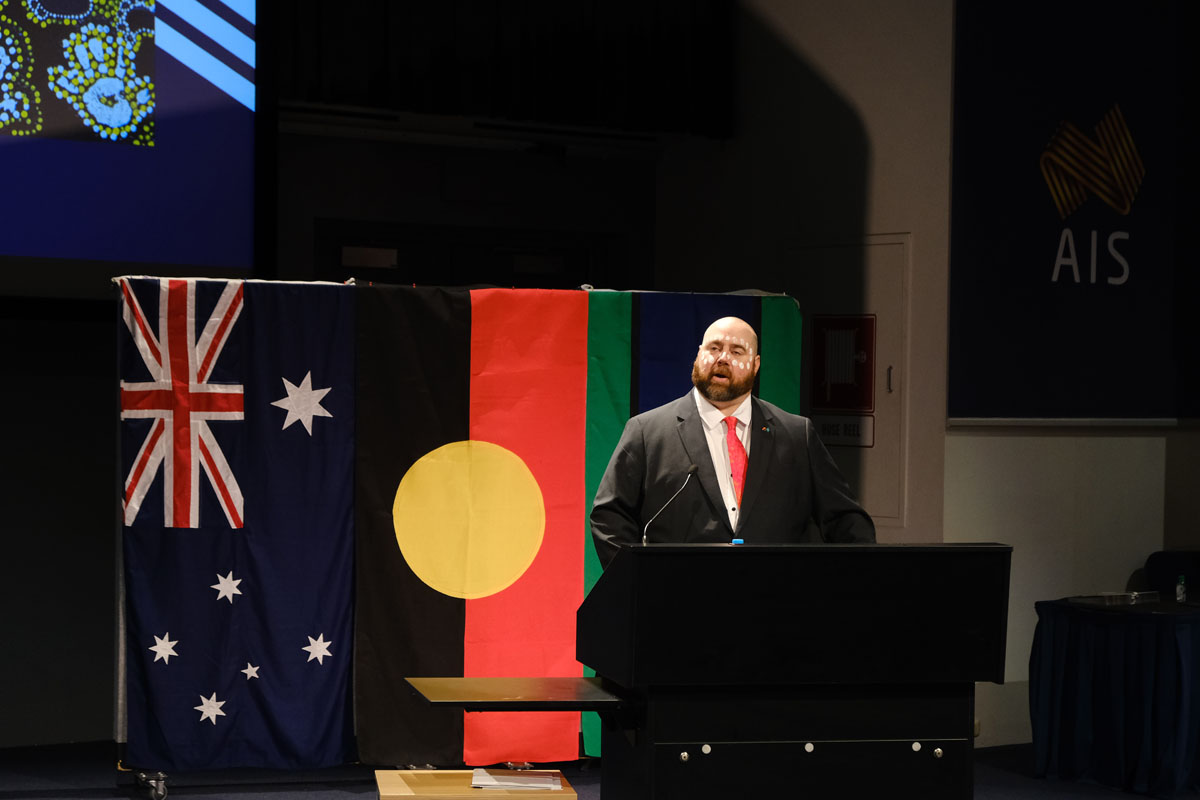 Erik Wilson stands behind a lecturn, wearing traditional facepaint around his eyes. He is standing in front of Australian, Aboriginal and Torres Strait Island flags.