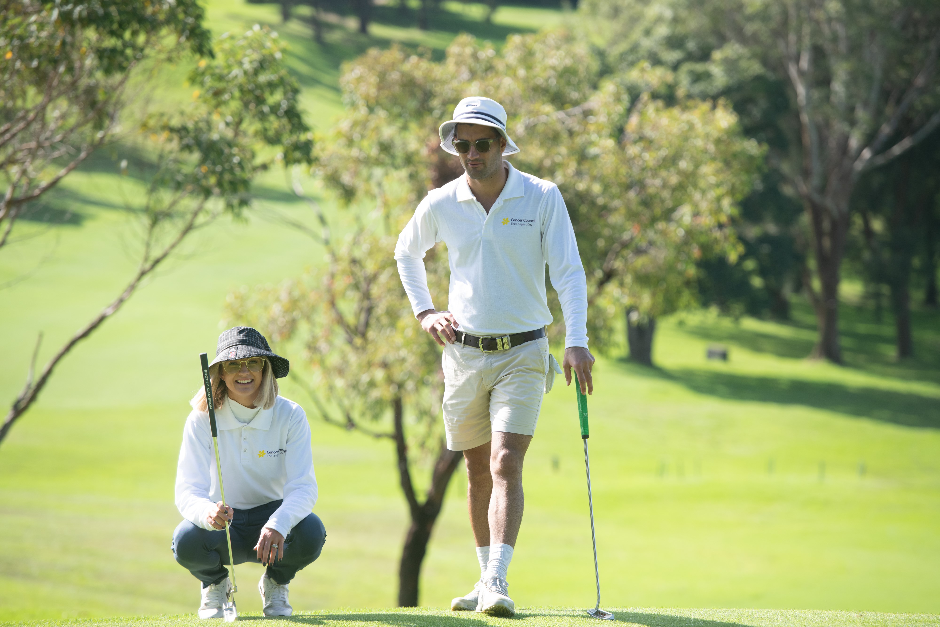 A woman and a man on a golf course holding golf clubs wearing wide brim hats and Cancer Council long sleeved shirts.
