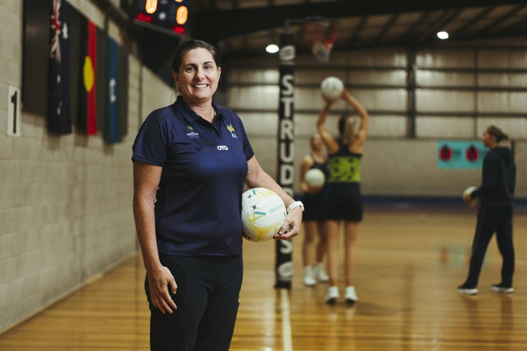 Kylee Byrne stands holding a netball with netball players shooting behind her.