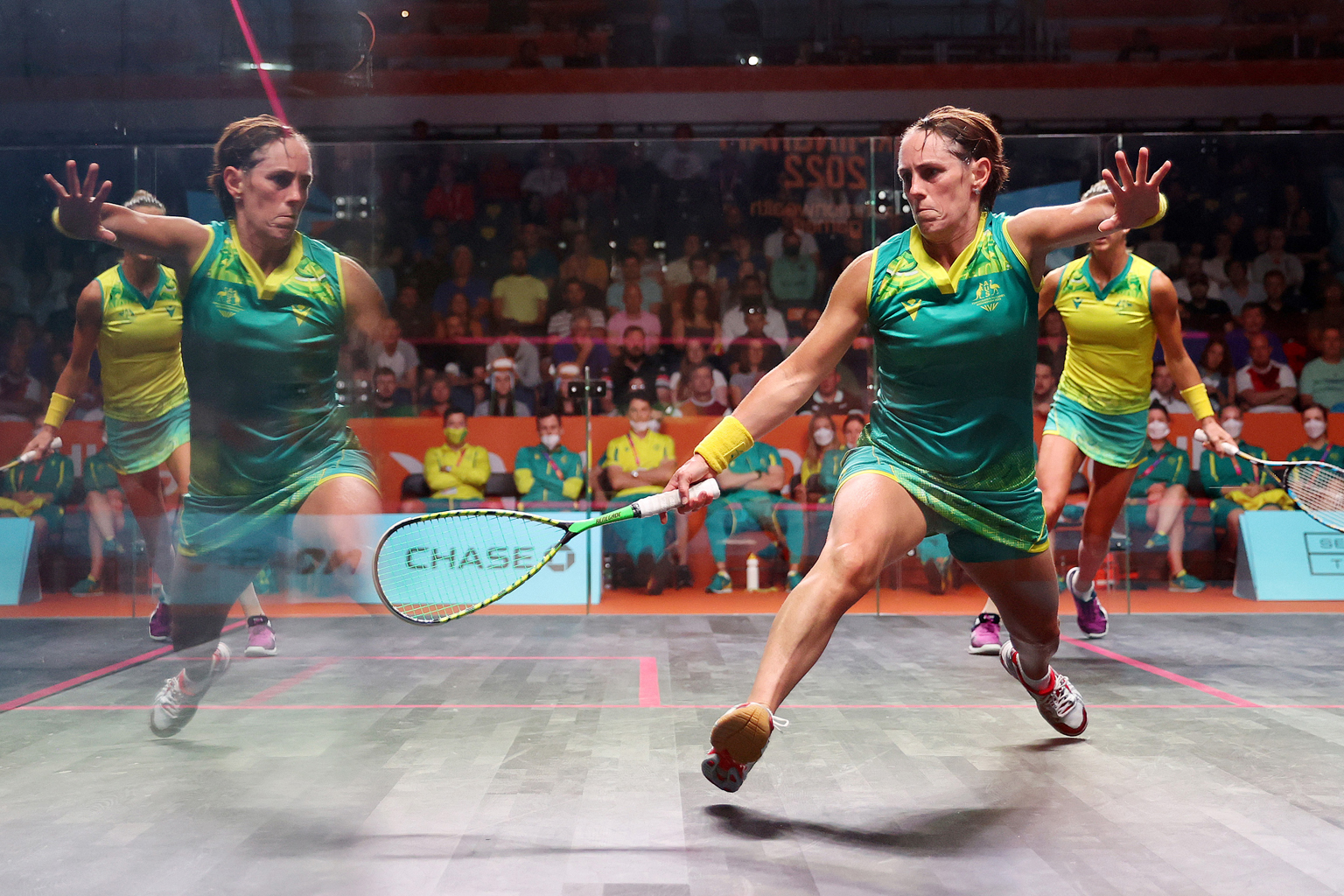 A squash player reaches for the ball, with her reflection clearly visible in the court's perspex wall.