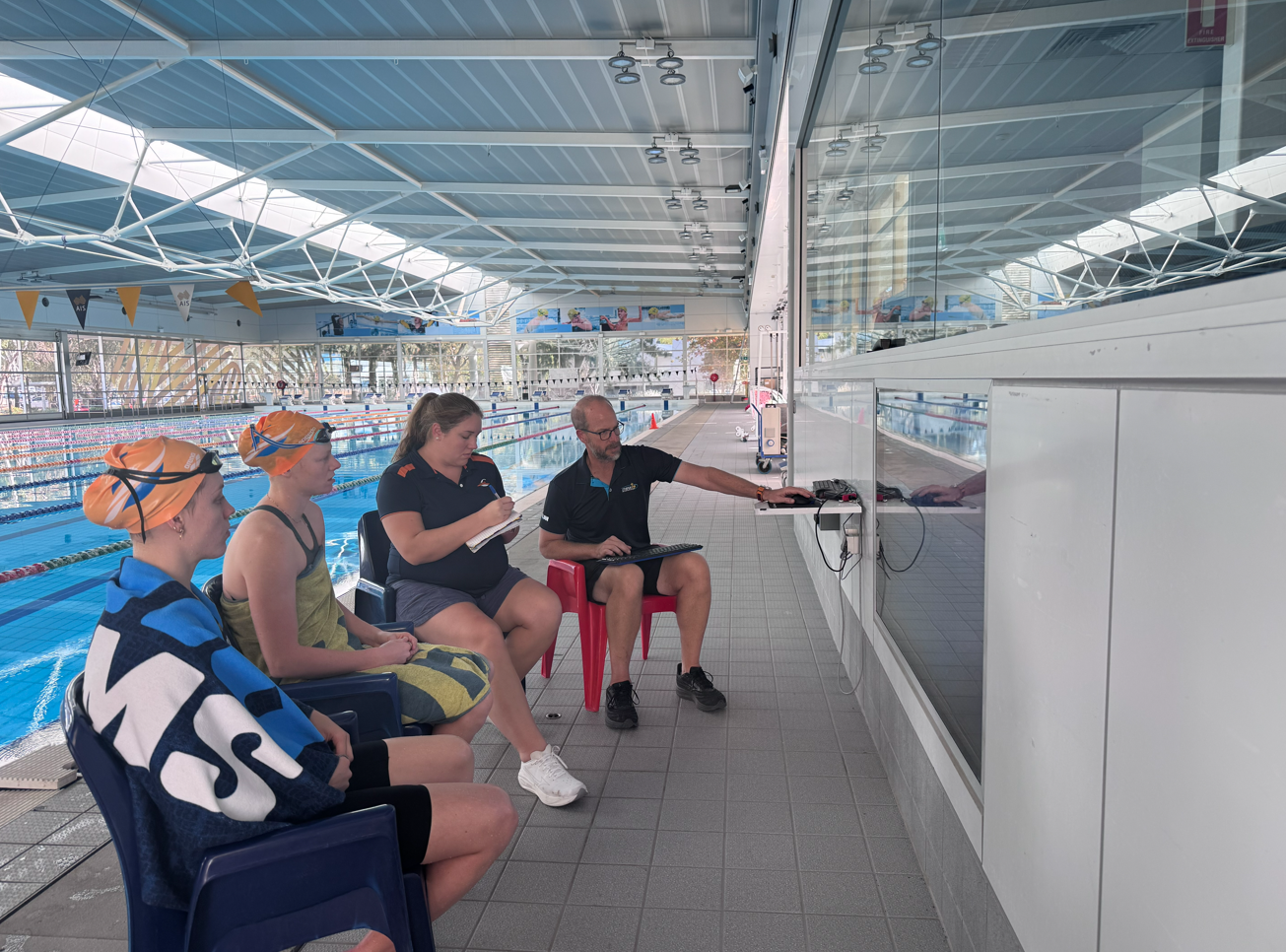 four people sitting on the side of an olympic pool viewing data on a large screen
