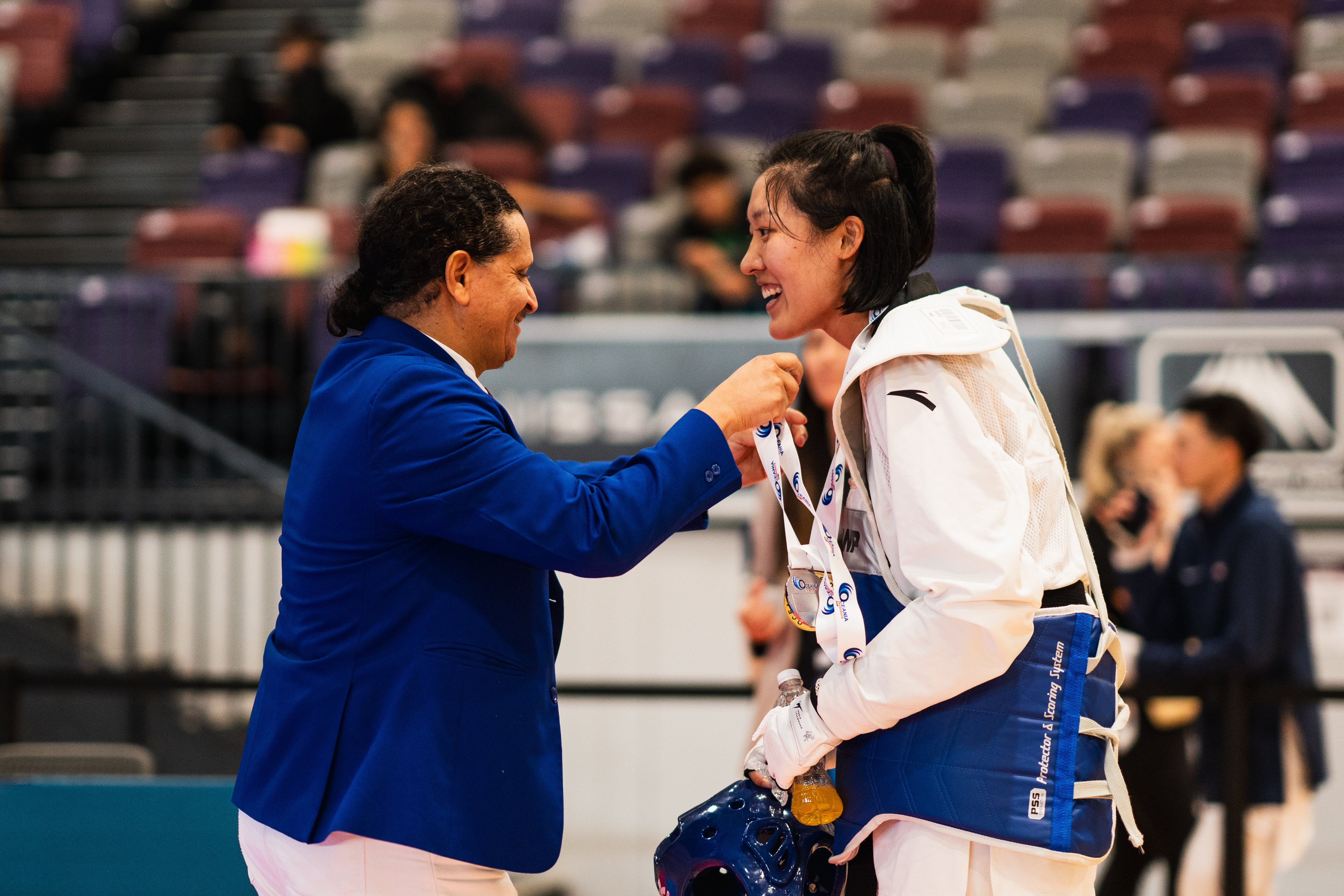 A Taekwondo official wearing a blue jacket places a medal around the neck of a young athlete who is wearing a white dobok. They are both smiling.