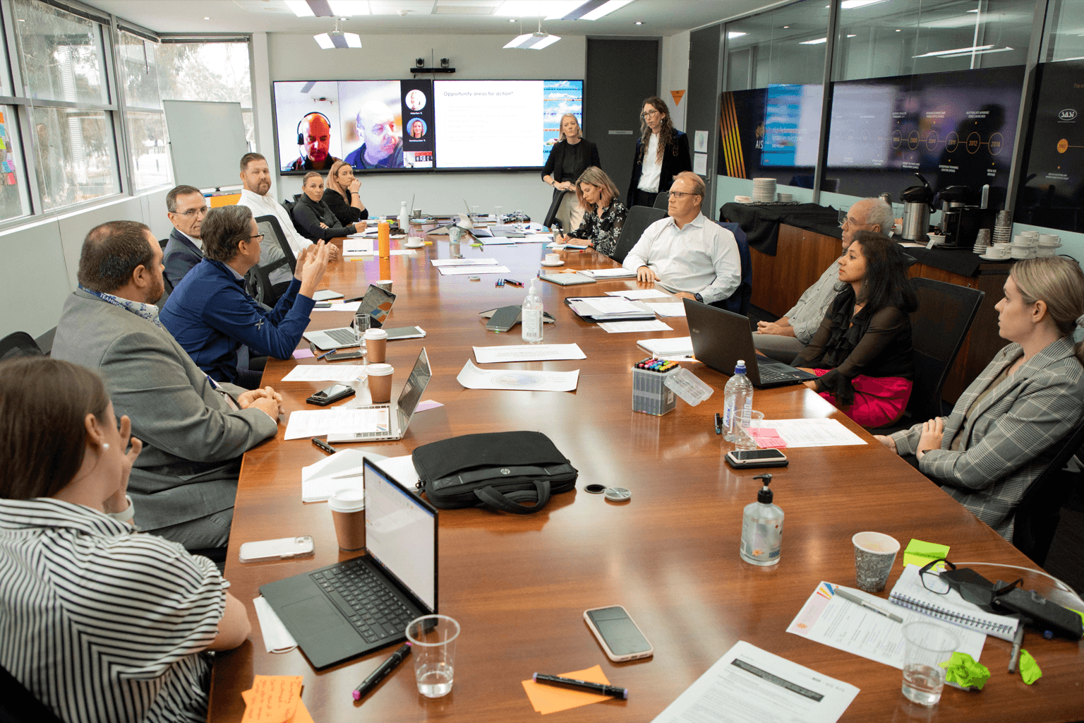 A group of people sit gathered around a boardroom table with large screens at one end.