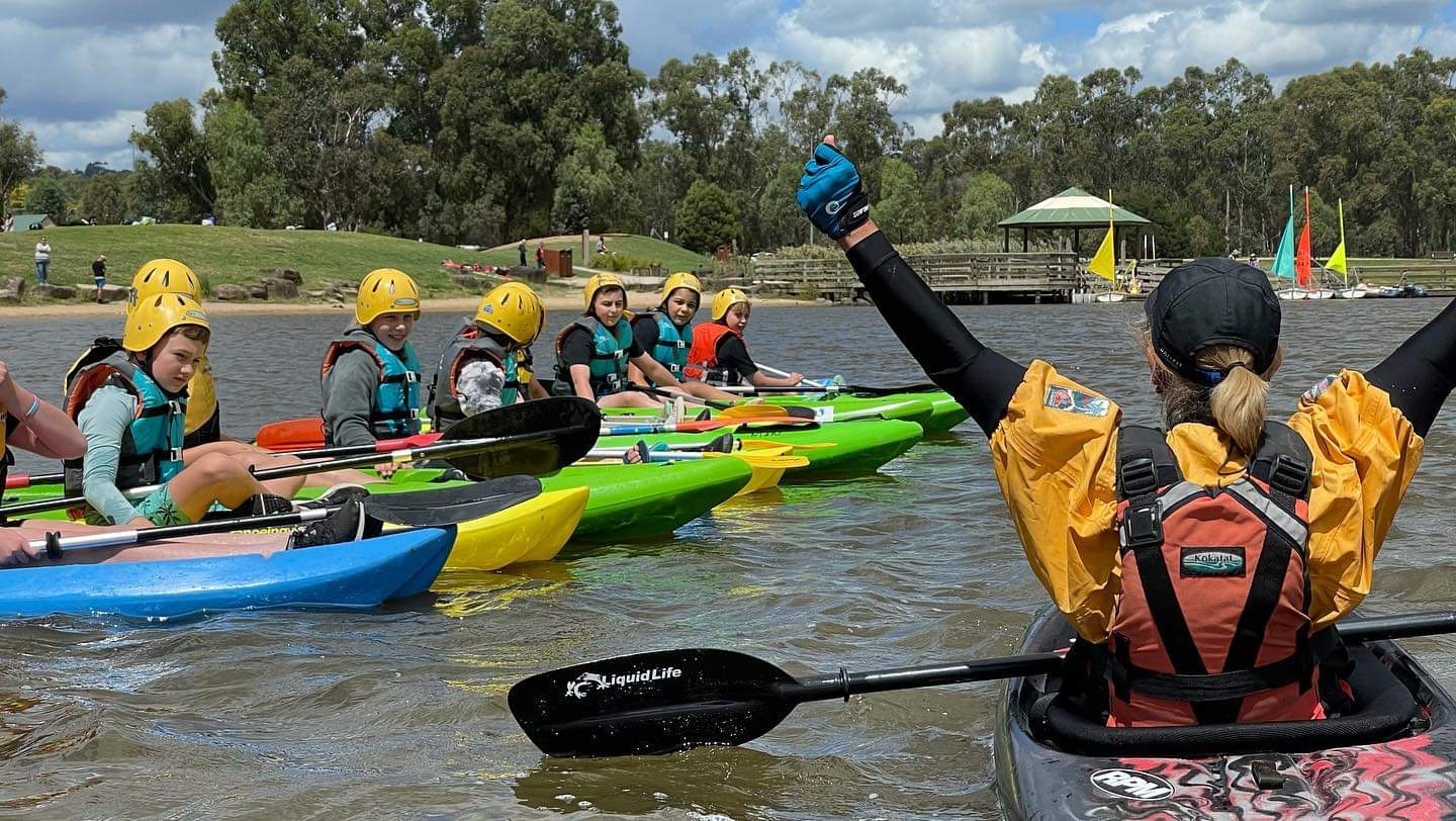 a group of students learning to paddle in a lake with a coach surrounded by lush green trees