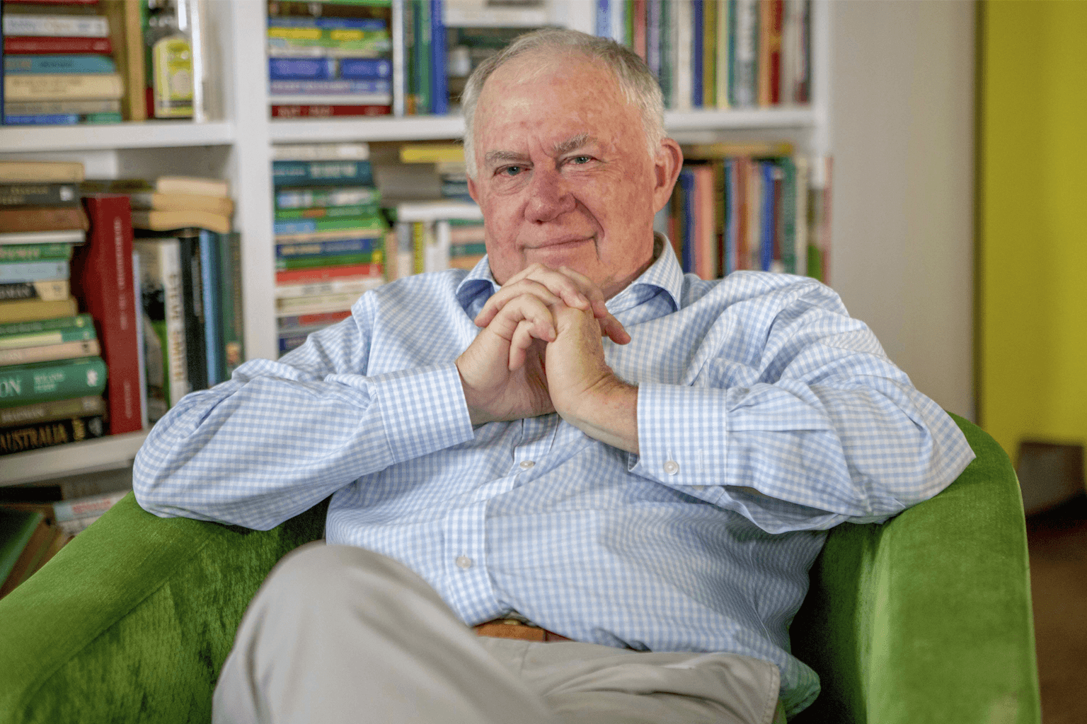 ABC Broadcaster Jim Maxwell sits in a green couch in front of a crowded bookcase in his Sydney home.