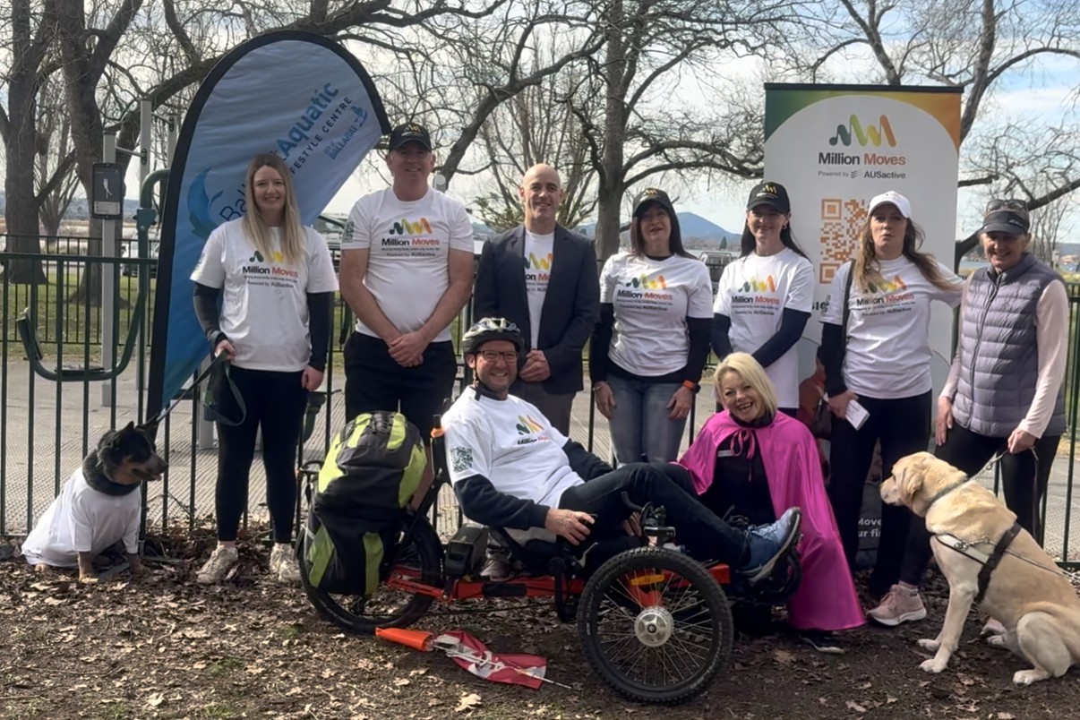 Nine people celebrate the launch of AUSActive's Million Moves initiative in Ballarat at a community space, accompanied by two assistance dogs