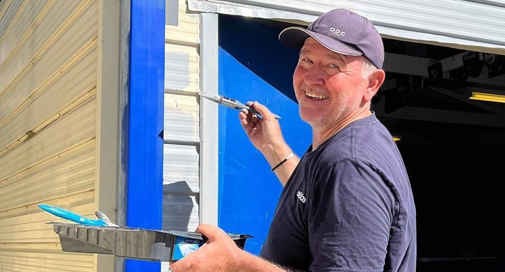 A male volunteer wearing a navy tshirt and cap smiles at the camera, he is holding a brush in one hand and a tray in the other as he paints a storage shed