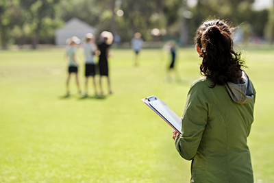 A female coach with a clipboard watching the team practicing on sports field