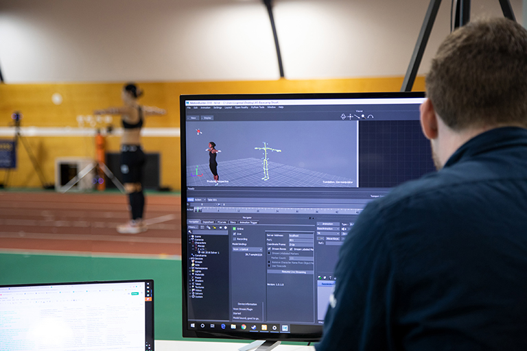 Image of a biomechanist analysing data at a computer of an athlete who is pictured in the background