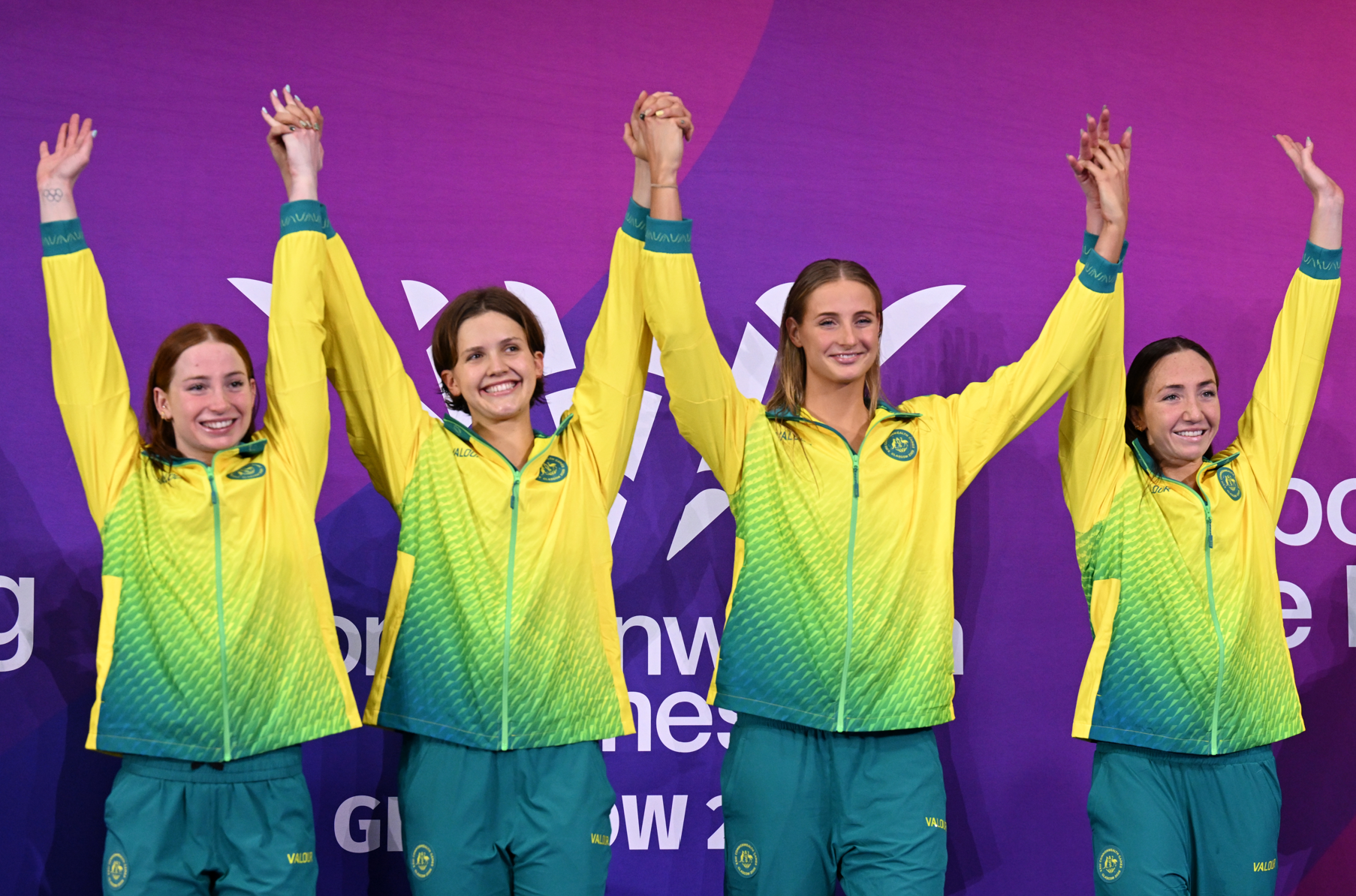 Four female swimmers with arms raised while standing on the medal dais in green and gold uniforms