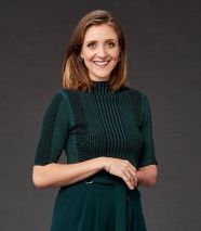 Smiling lady with brown hair. Wearing a dark green dress, standing against a dark grey background